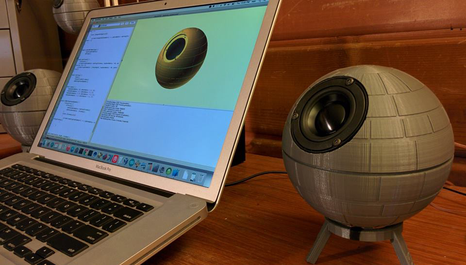Shows a laptop running OpenSCAD with the design for the speaker housing that looks like the Deathstar from the Star Wars franchise with 2 of such speakers either side of the user's laptop.